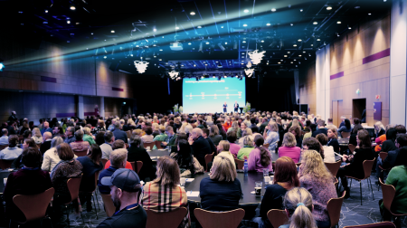 Fullsatt sal som følger med på presentasjon på storskjerm. En lyskaster kaster stripene i DFØ-logoen over forsamlingen.